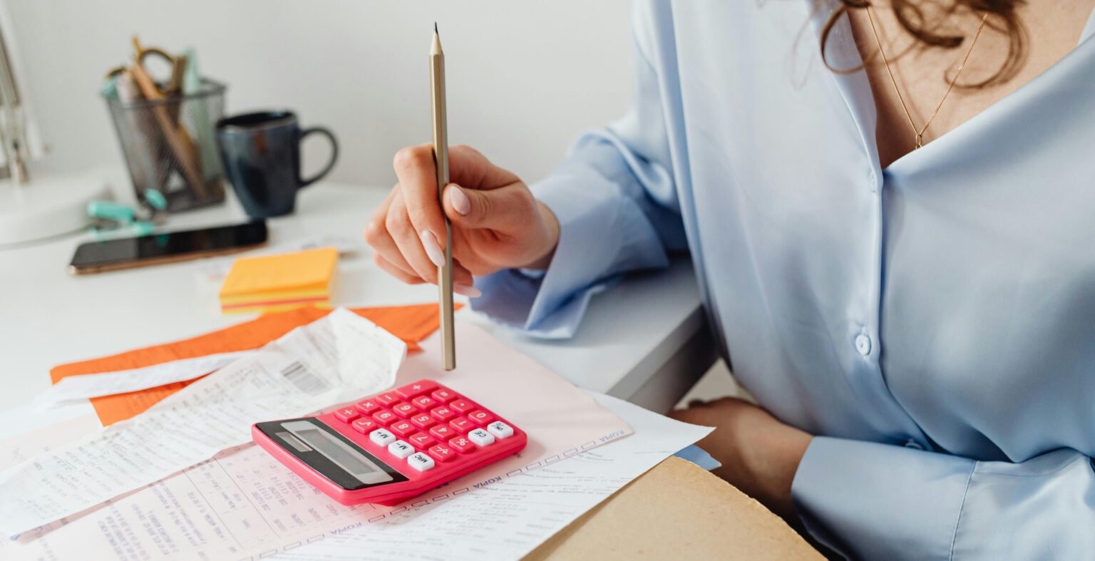 Comment créer un budget efficace : conseils, outil budget planner annuel automatisé. Image où on voit une femme tenir un crayon à papier dans sa main droite qui est posée sur son bureau. Sur son bureau elle a des documents papier et une calculatrice.